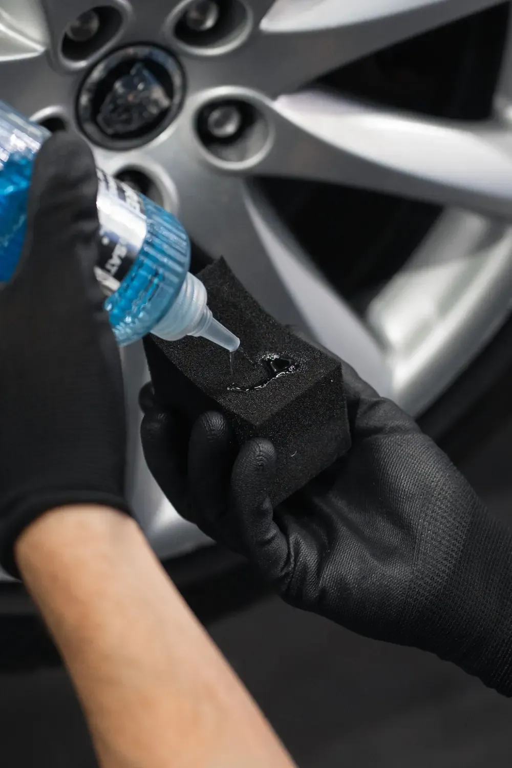 Un flacon bleu verse du produit sur un applicateur en mousse noire, devant une jante